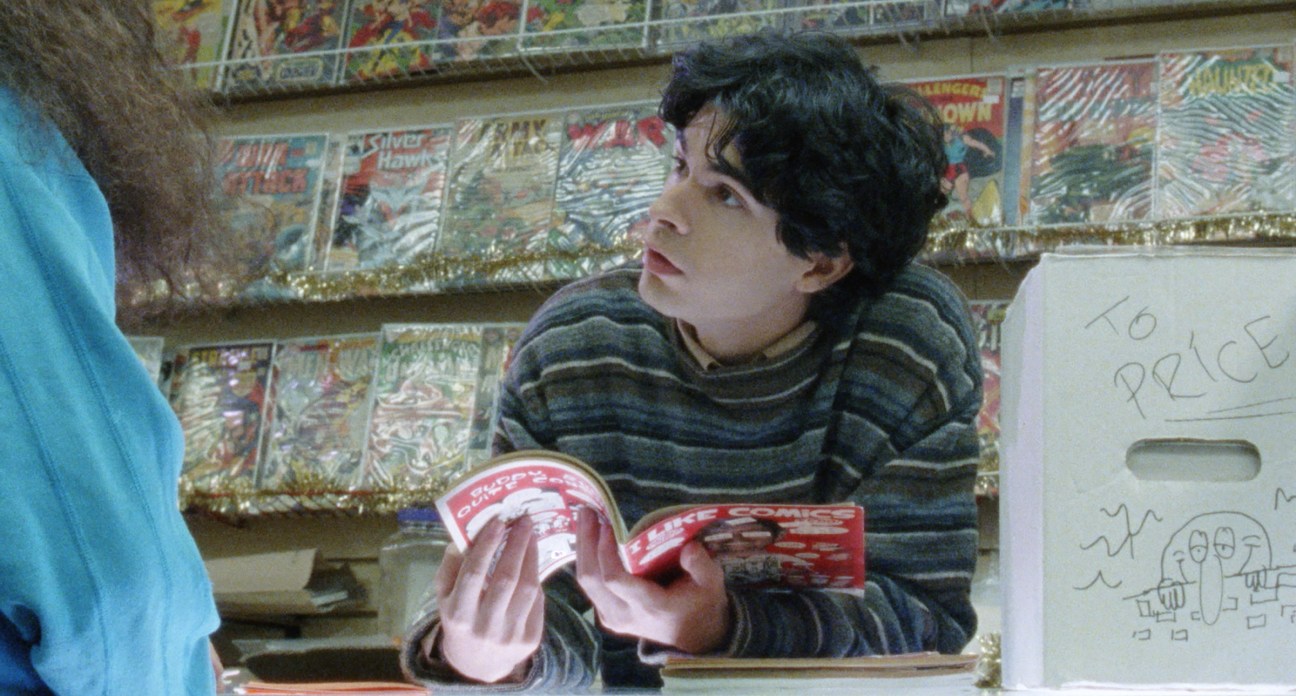 Young man with dark curly hair reading comic books in a store surrounded by comic book displays