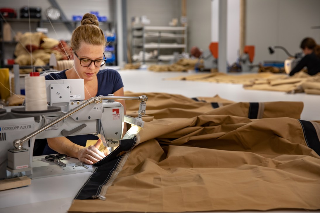 A worker sews tan fabric on an industrial sewing machine in a manufacturing facility.