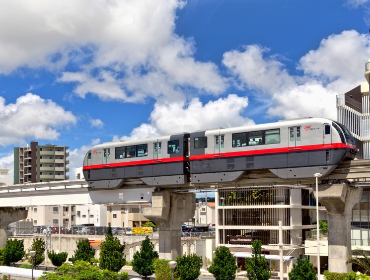 A red and white monorail train glides along an elevated track above a urban cityscape under a bright blue sky.