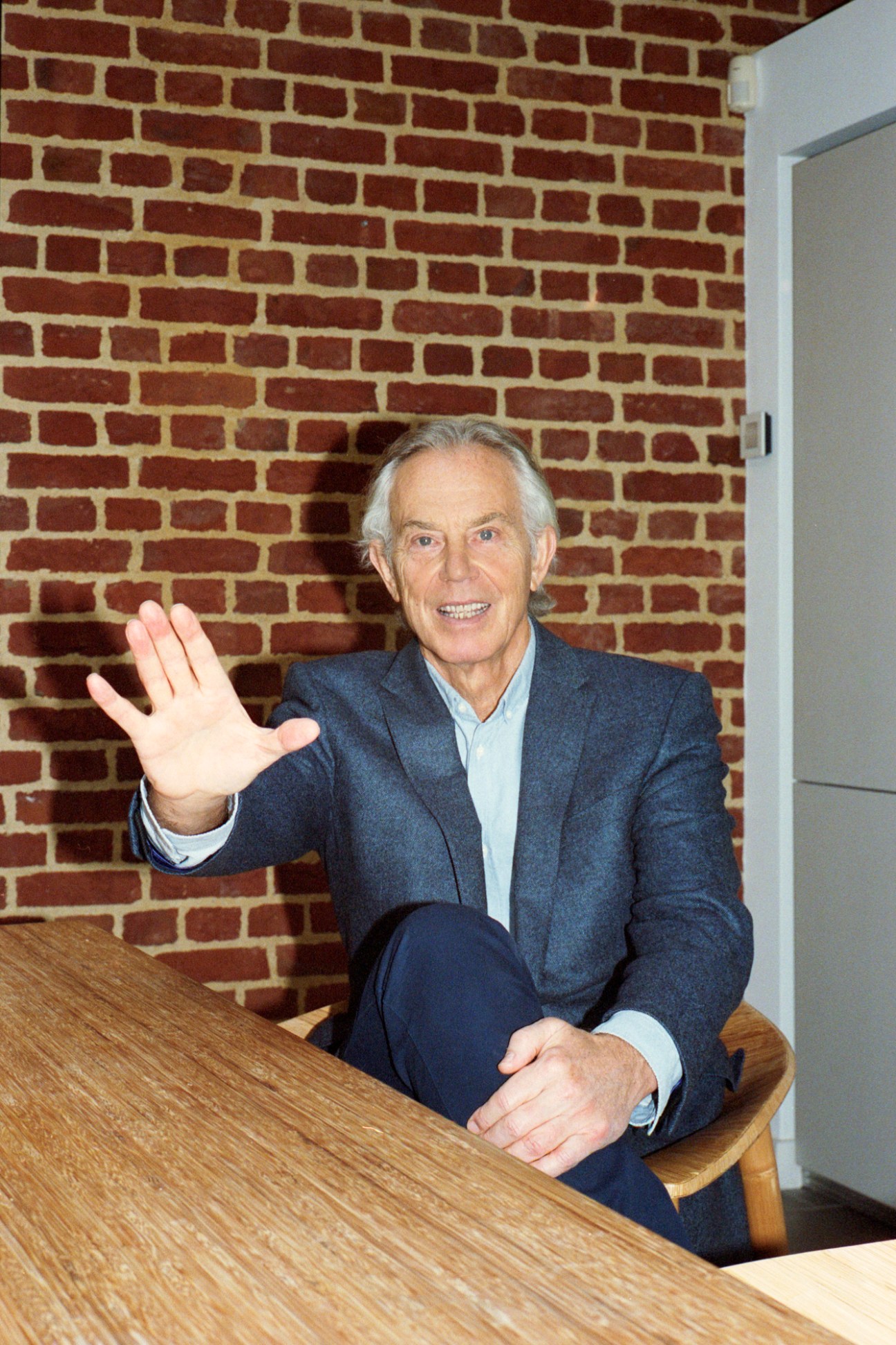 Former UK Prime Minister Tony Blair waving while seated at a wooden table in an office setting