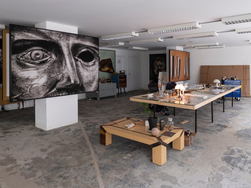 Artist's studio with large portrait artwork on wall, wooden tables, lamps, and art materials in concrete space