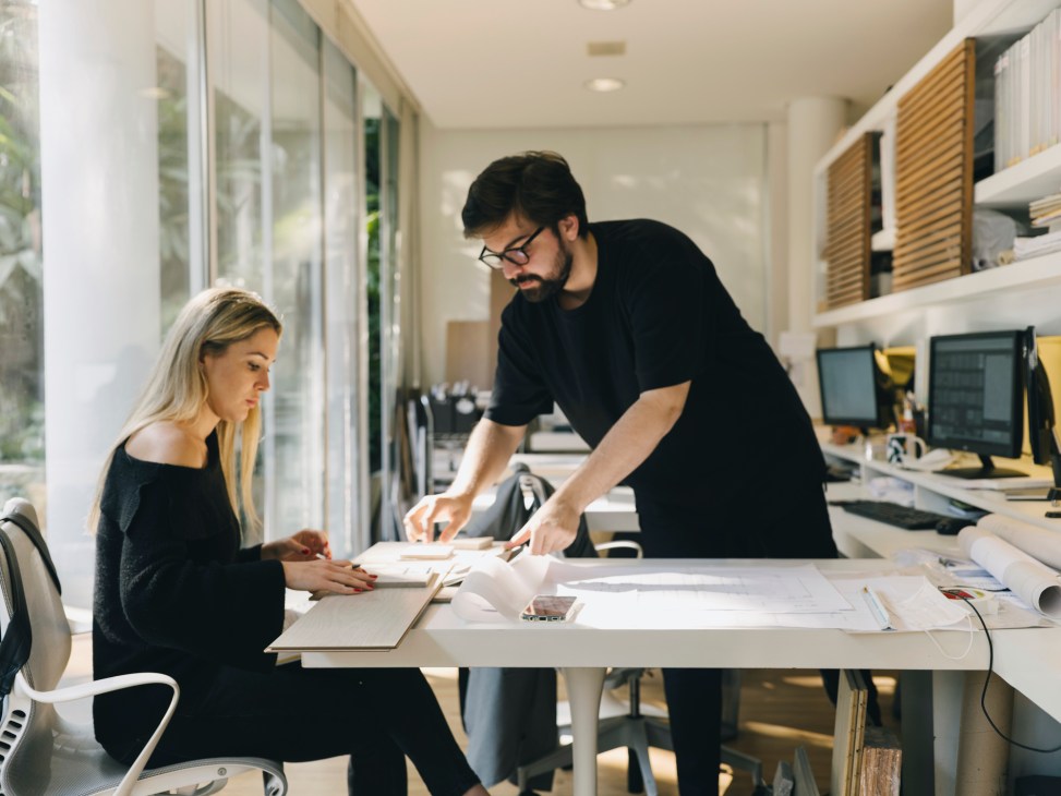 Two architects reviewing architectural drawings and plans spread across a white desk in a modern office