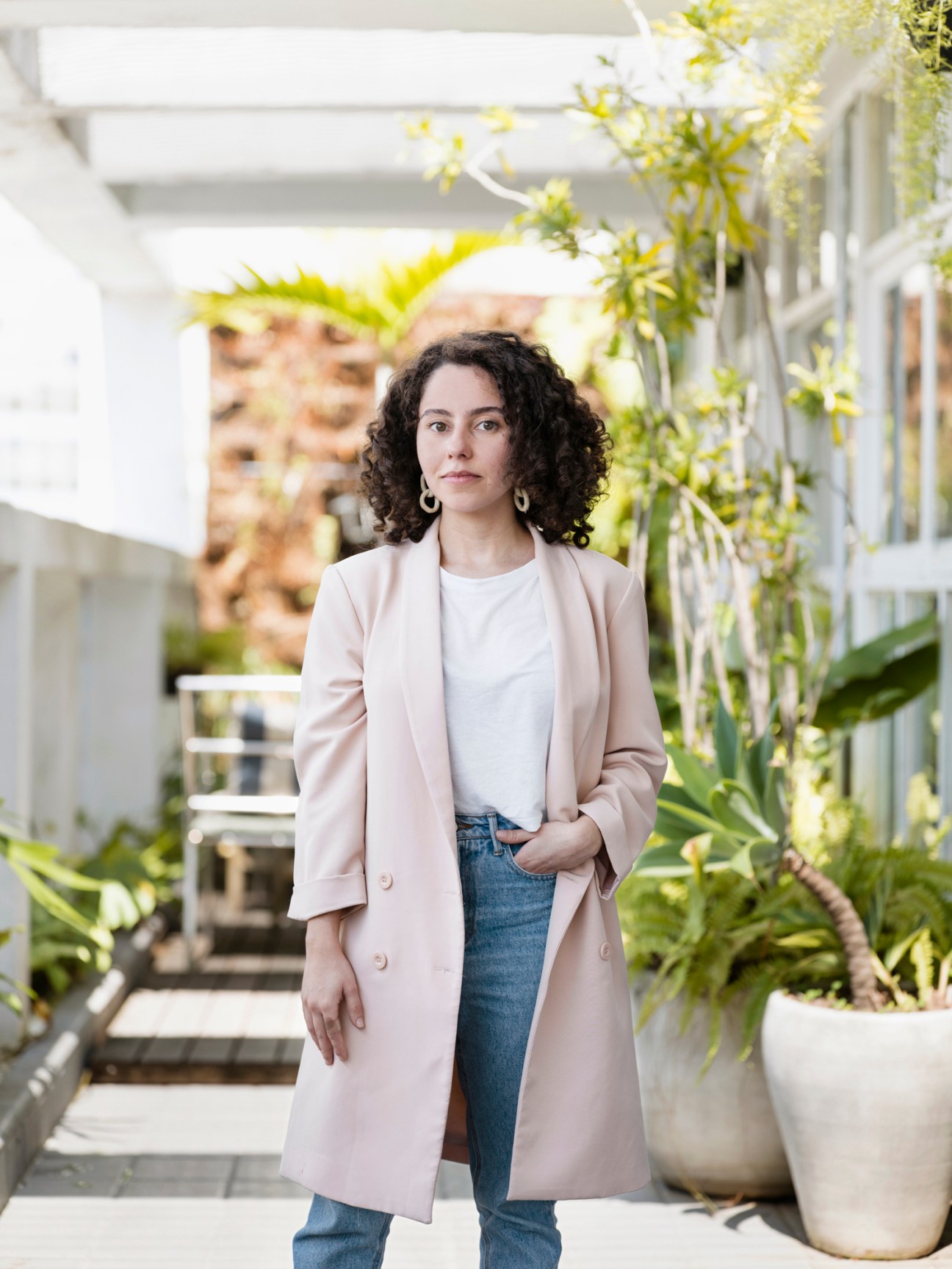 Woman with curly dark hair wearing light pink blazer and white top standing in modern courtyard with plants