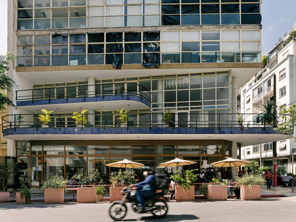 Modern building with glass facade, outdoor seating area with umbrellas and planters on street level