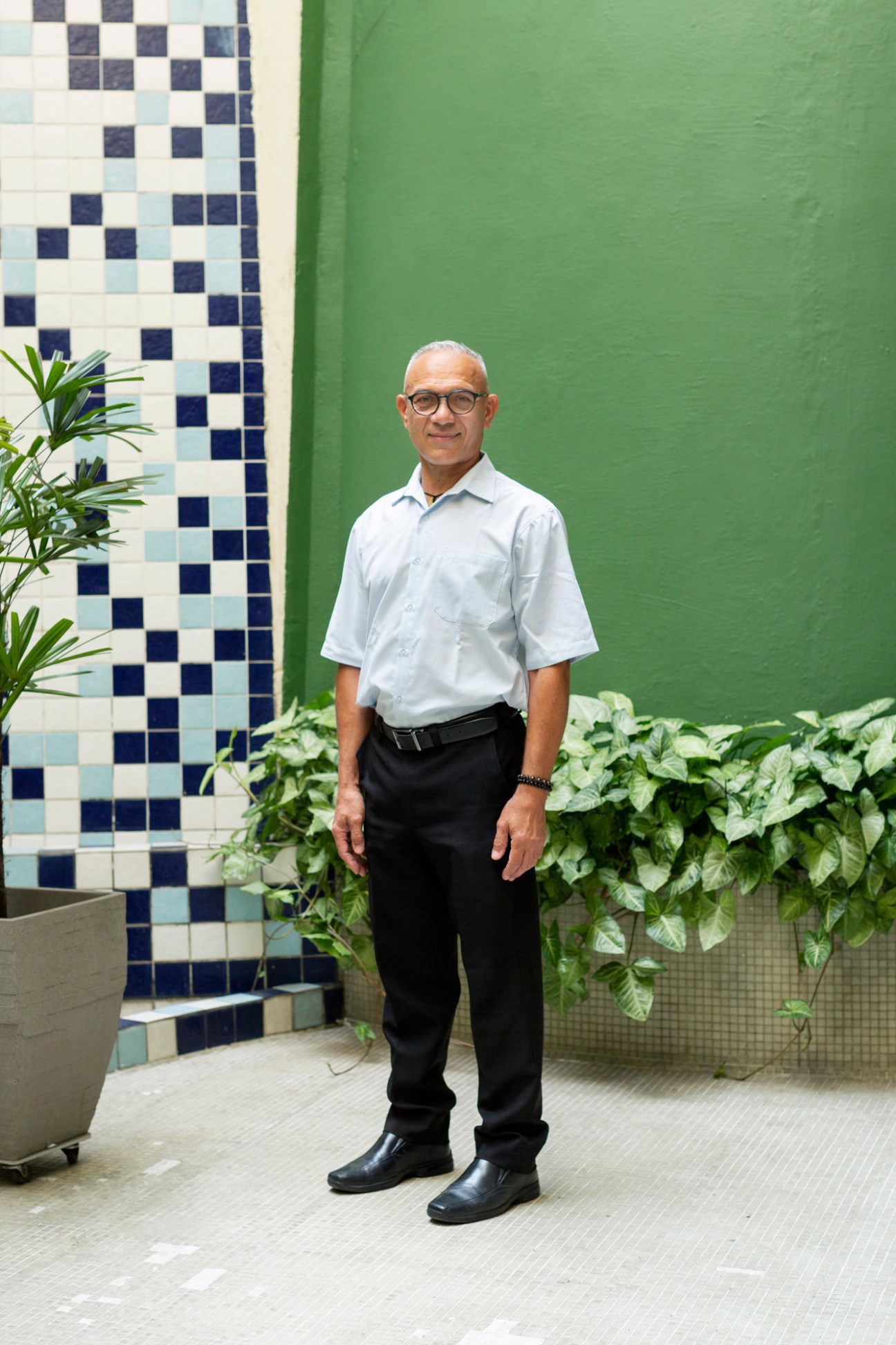 Man in glasses wearing white shirt and dark pants standing in front of green wall with plants and mosaic tiles