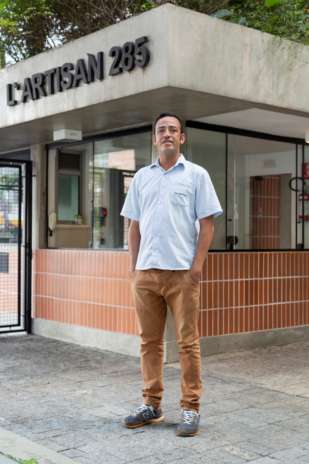 Man in white polo shirt and brown pants standing outside modern building with L'ARTISAN 285 signage