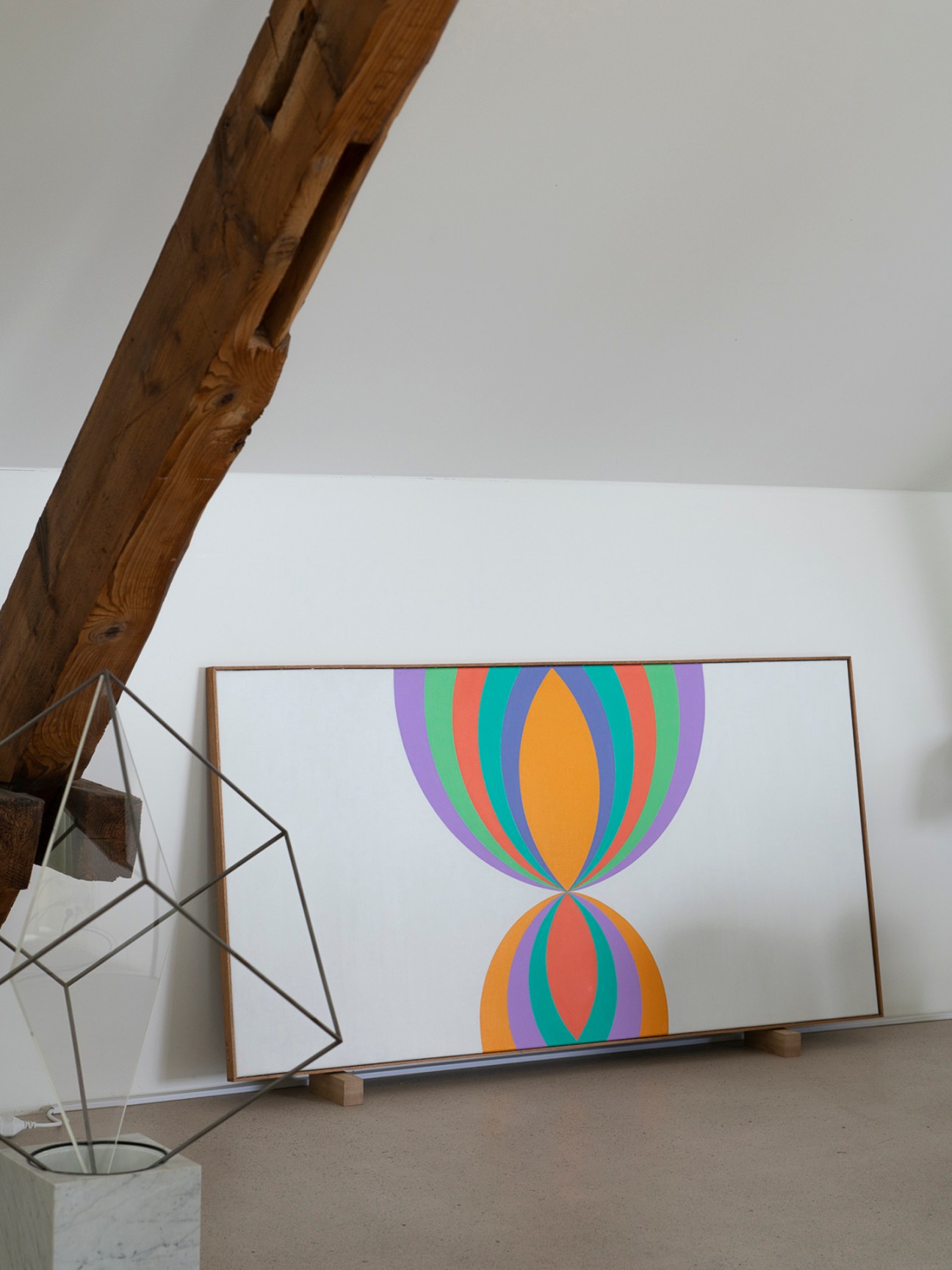 Framed colorful geometric artwork featuring interlocking circles in rainbow stripes displayed in a minimalist gallery space with exposed wooden beam and potted plant.