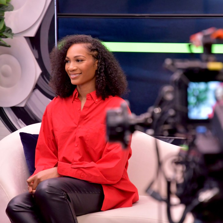 Woman in red shirt sitting on white chair in television studio with cameras in background