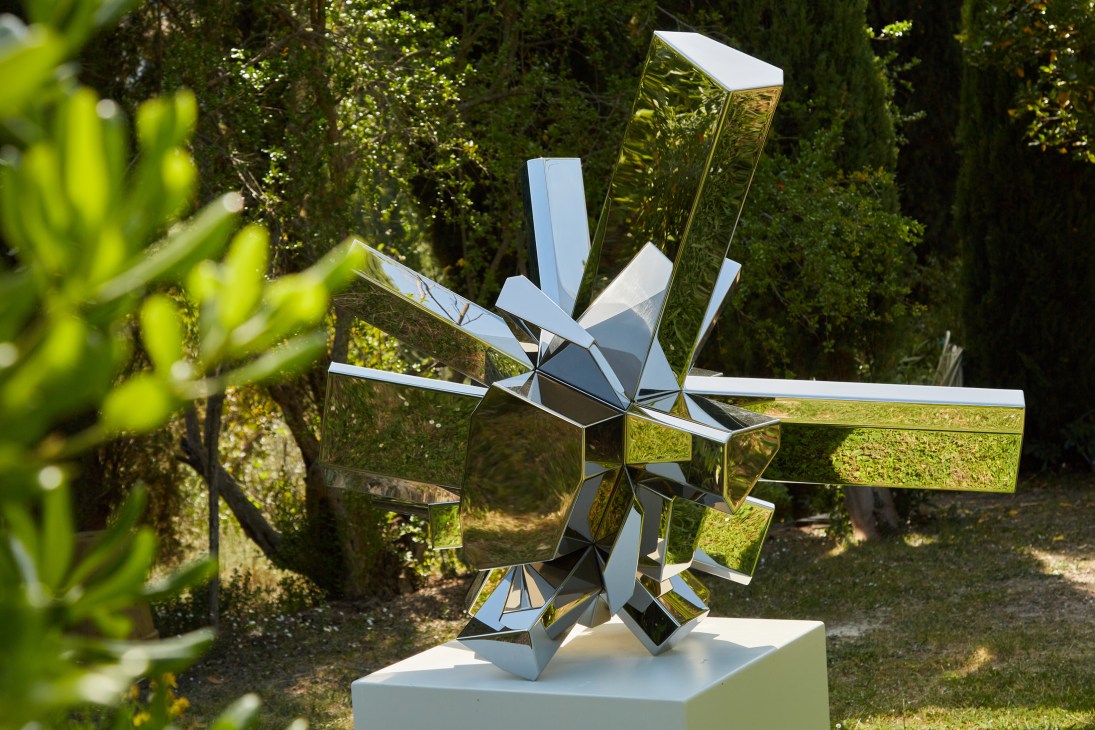 Reflective geometric metal sculpture with angular mirrored surfaces on white pedestal in garden setting