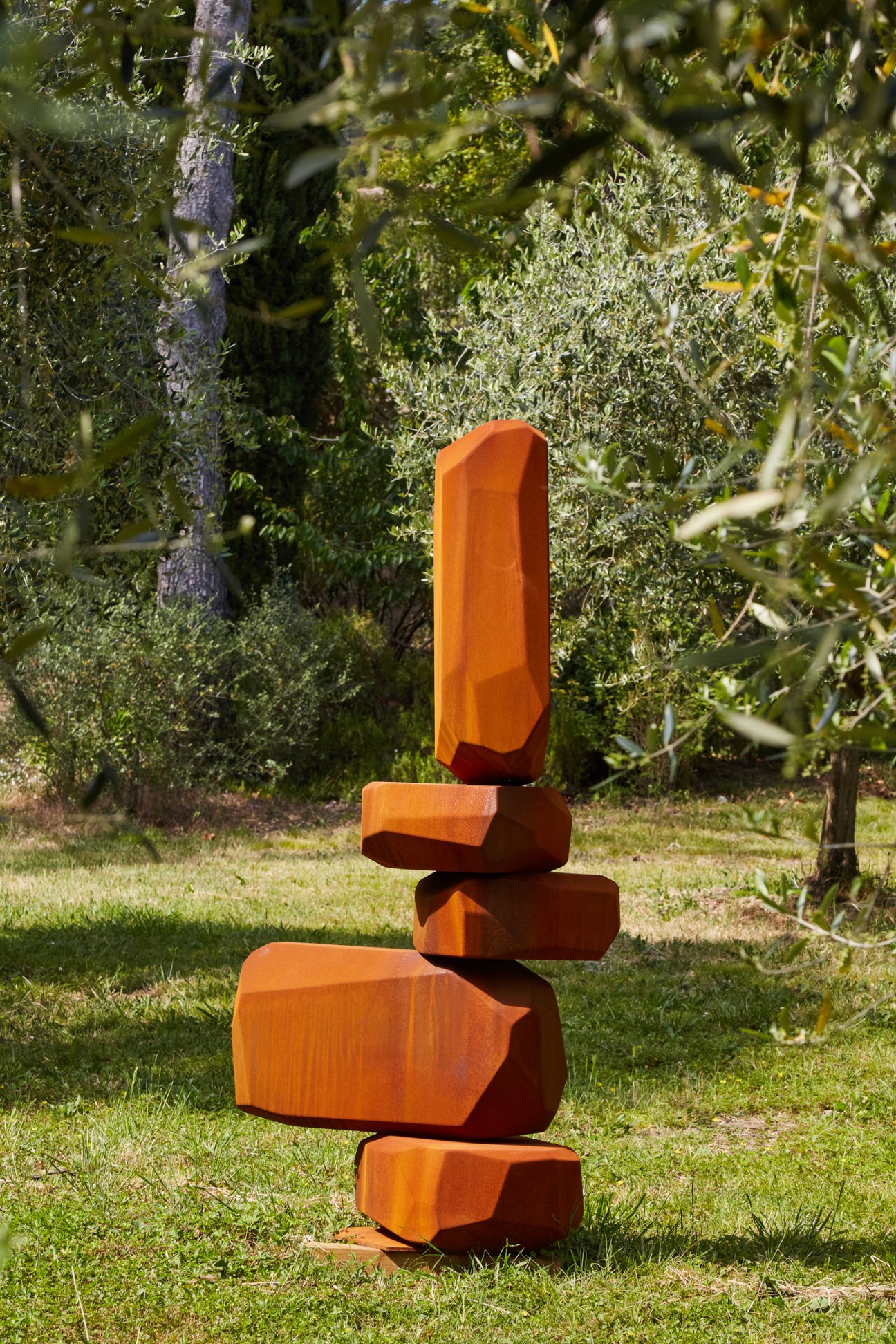 Orange geometric sculpture made of stacked angular blocks in a garden setting with trees in background