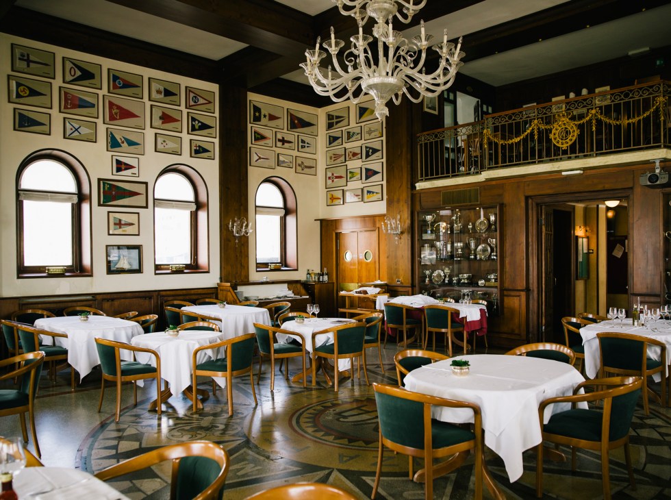 Elegant dining room with wooden chairs, white tablecloths, nautical flag collection, and ornate chandelier in Trieste.