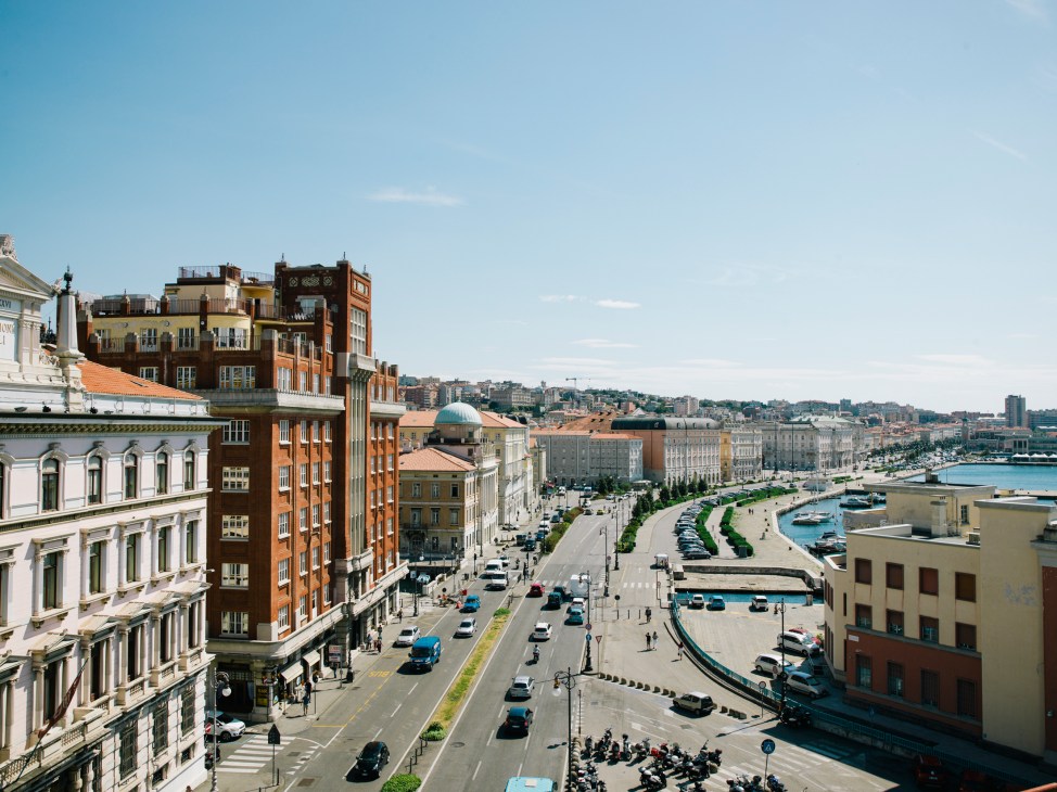 Elevated view of Trieste's waterfront with colorful historic buildings, a curved plaza, boats in the harbor, and hills in the background under blue sky.