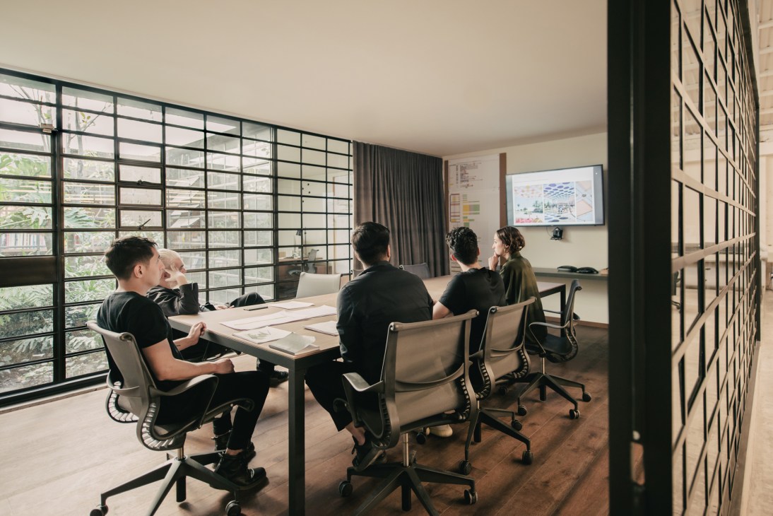 Four people sit at a wooden table in a bright meeting room with floor-to-ceiling grid windows overlooking greenery at Esrawe Studio.