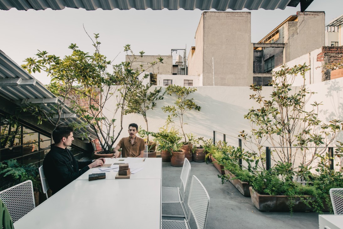 Two people sitting at a white table on a rooftop terrace surrounded by potted plants and greenery overlooking Mexico City buildings.