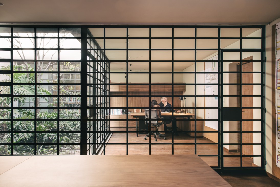 Interior of Esrawe Studio showing glass-panelled walls with black frames overlooking a garden, with a wooden desk workspace visible through the panels.