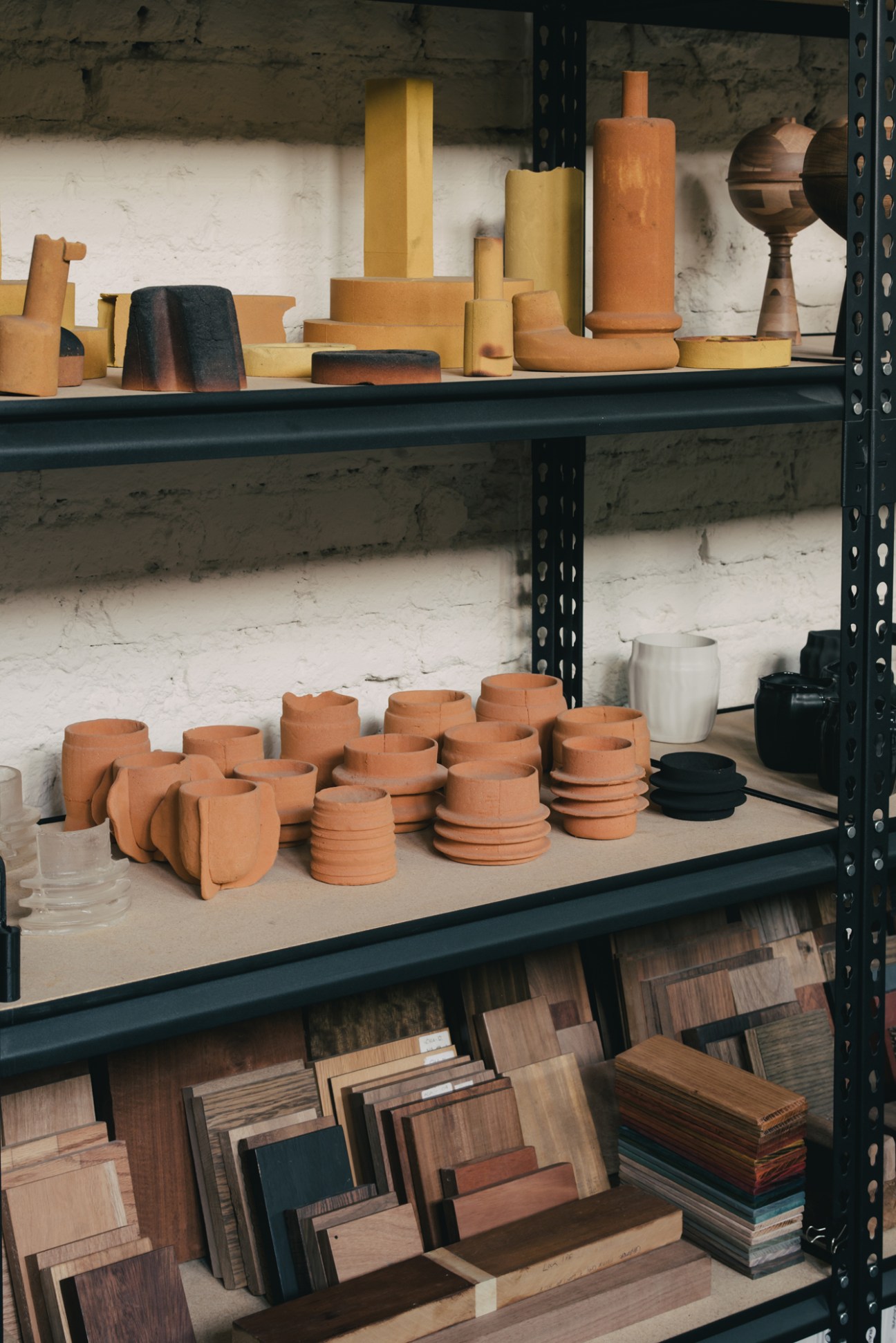 Wooden objects and terracotta vessels displayed on industrial metal shelving with white brick wall backdrop.