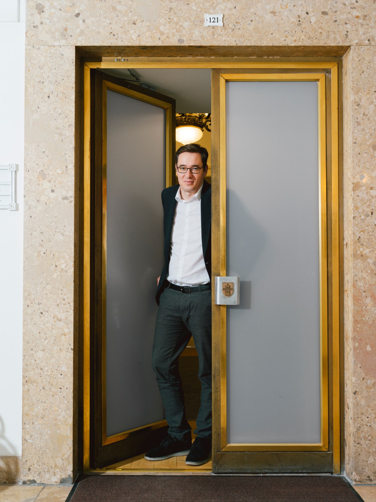 Gergely Karacsony standing in a modern elevator doorway wearing glasses, white shirt and dark jacket
