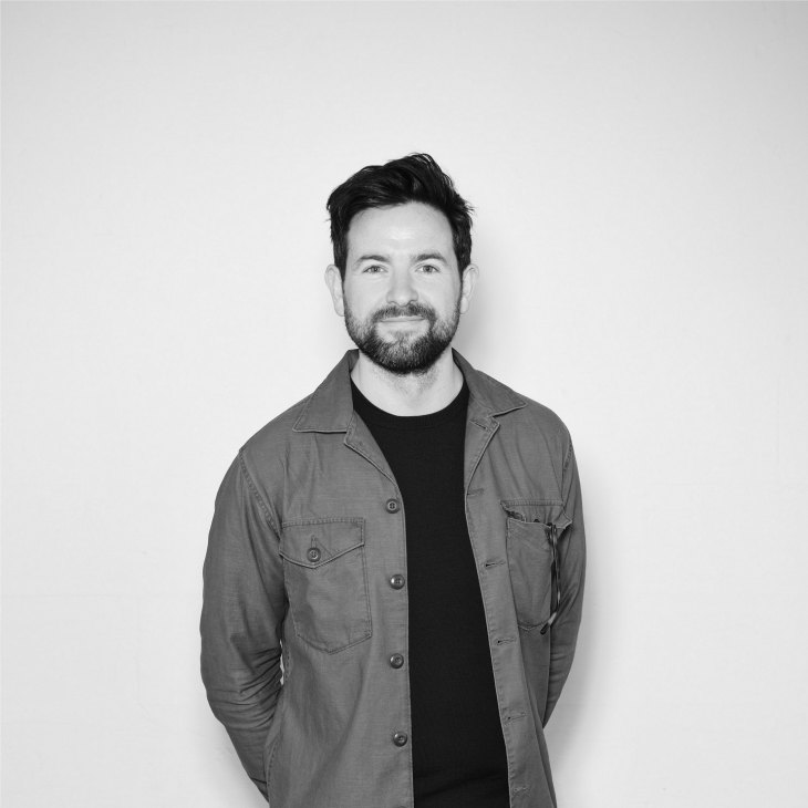 Black and white portrait of a man with dark hair and beard wearing a grey button-up shirt over a black t-shirt against a white background.