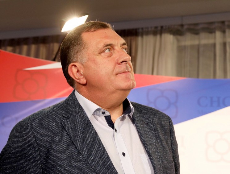 A man in a grey blazer looking upward, standing in front of a red, blue, and white flag backdrop at what appears to be a political event.