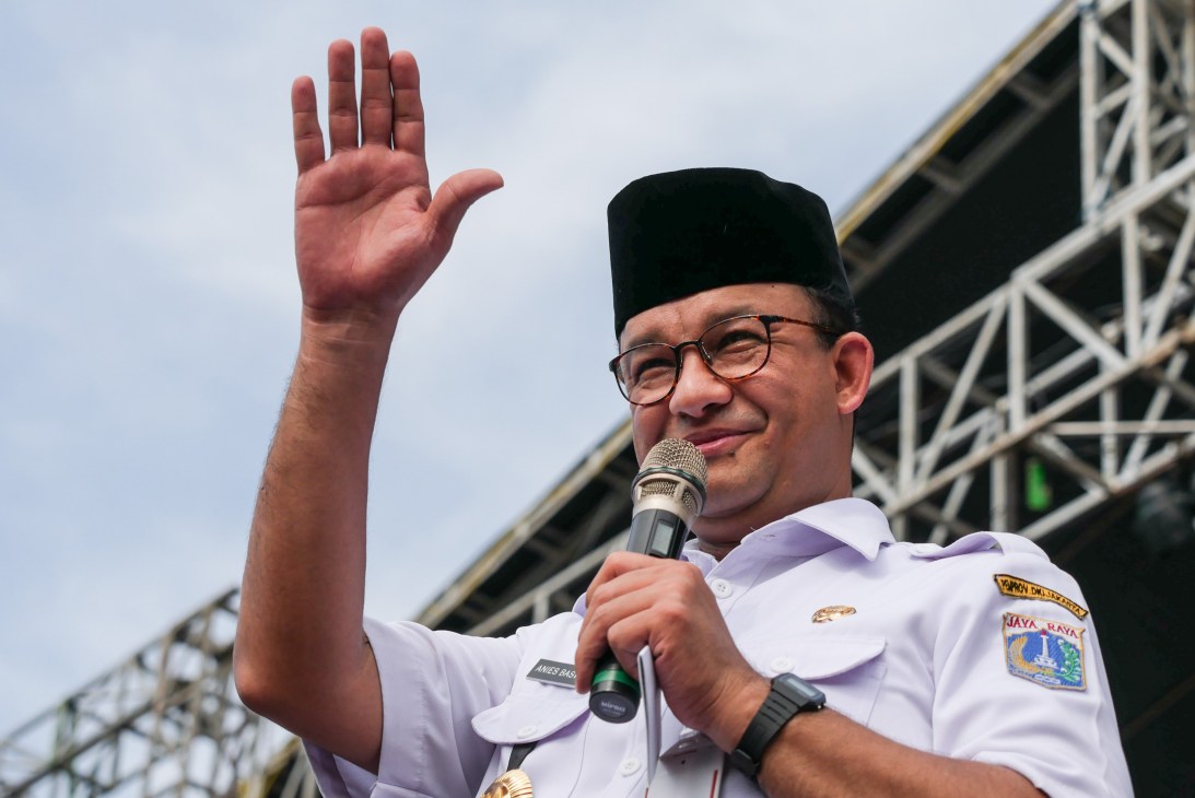 Anies Baswedan speaking at a political event with microphone, waving and wearing glasses and black cap
