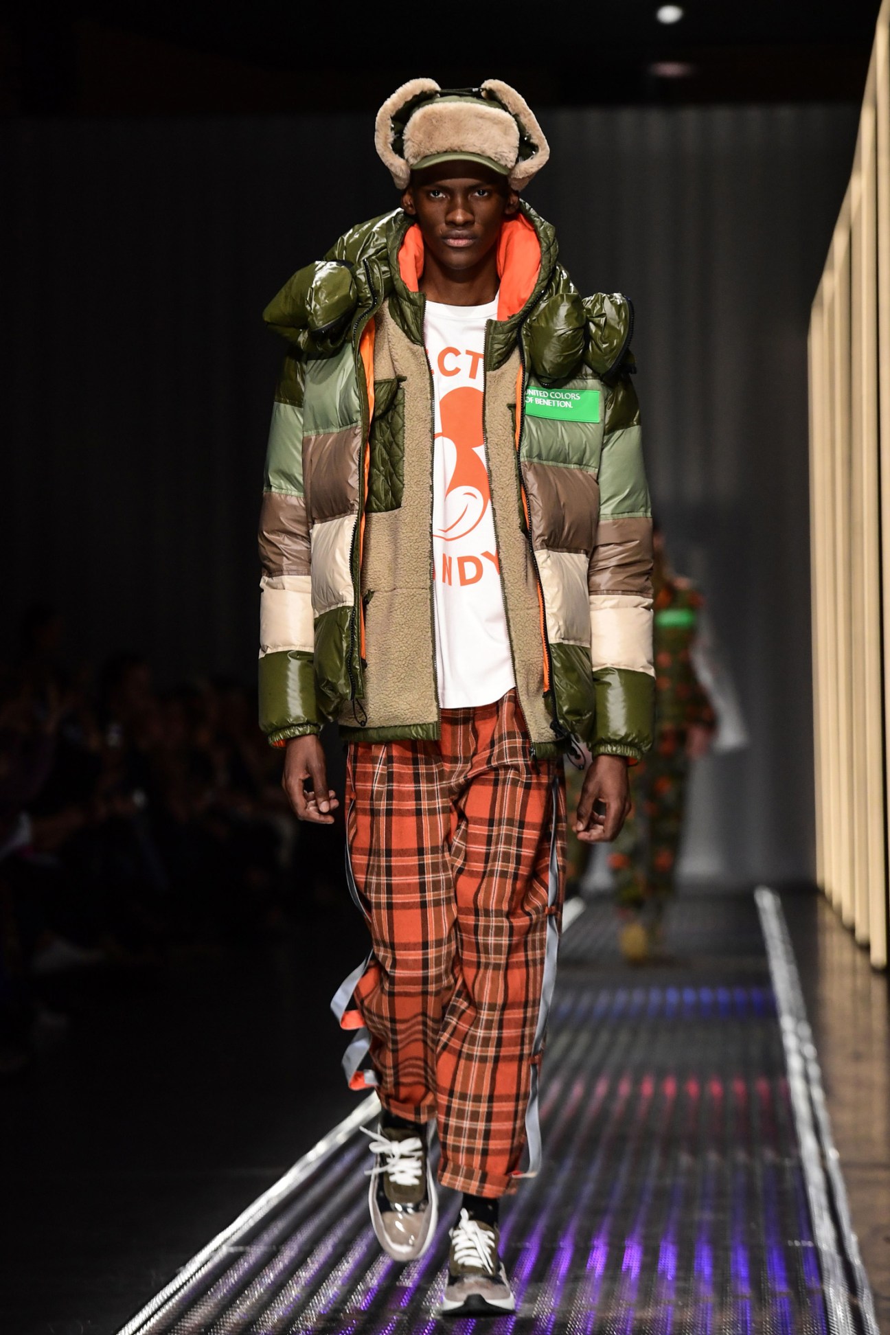 Model walks runway in Benetton color-block puffer jacket with plaid pants and oversized fuzzy hat.