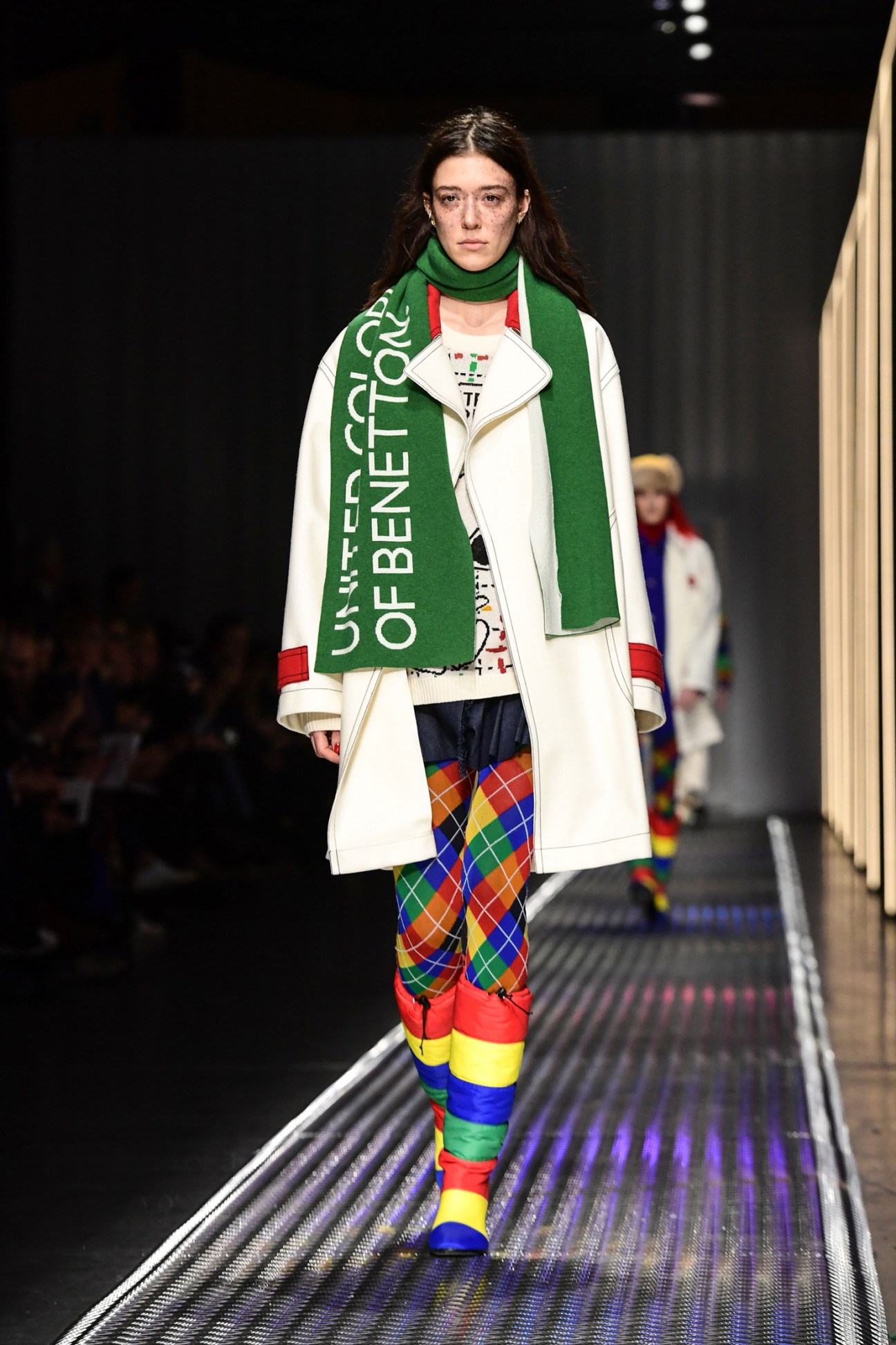 Model on runway wearing Benetton white coat with green scarf and rainbow argyle leggings.