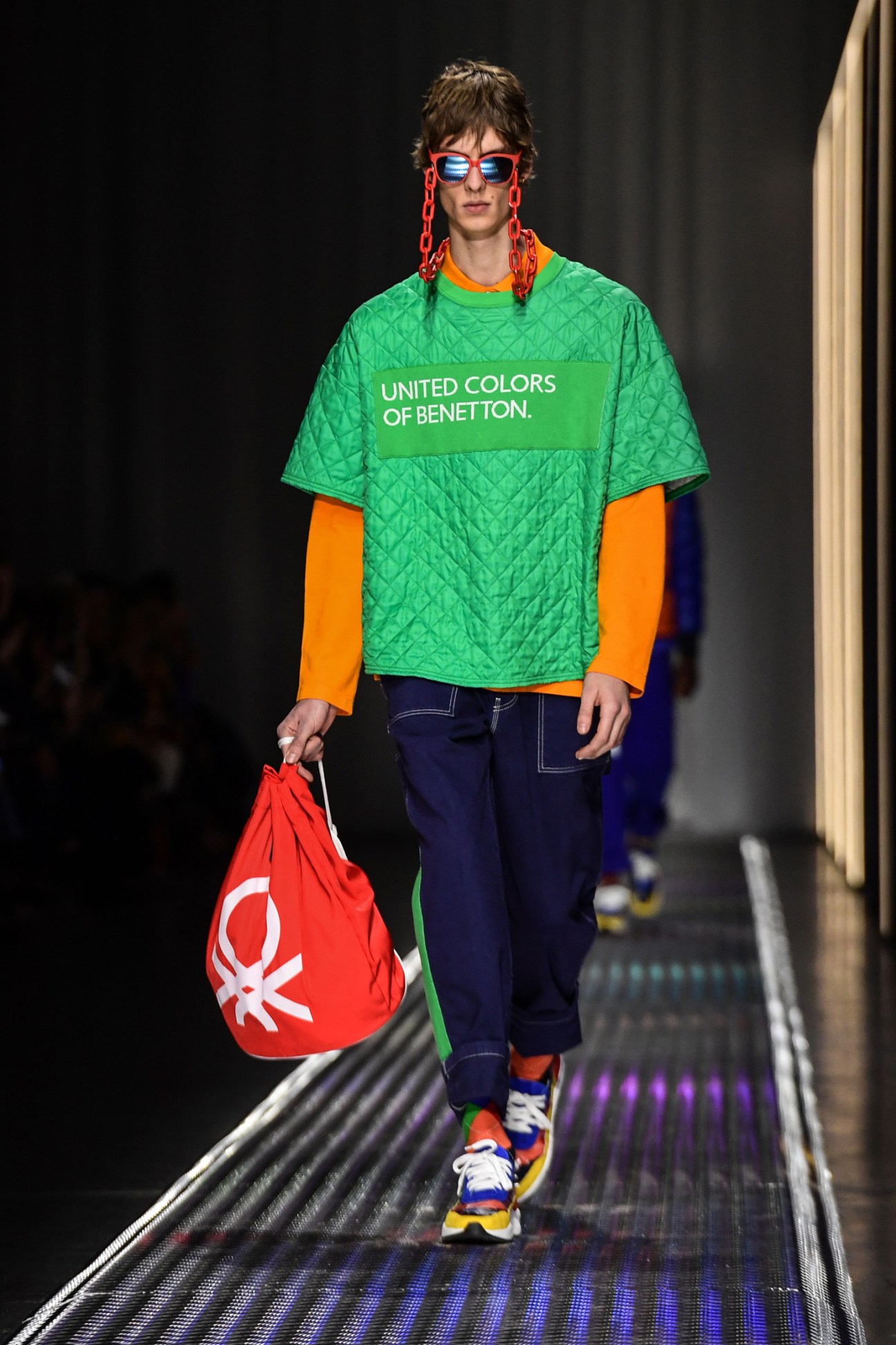 Model on runway wearing colorful Benetton outfit with green quilted shirt, orange sleeves, and red accessories.