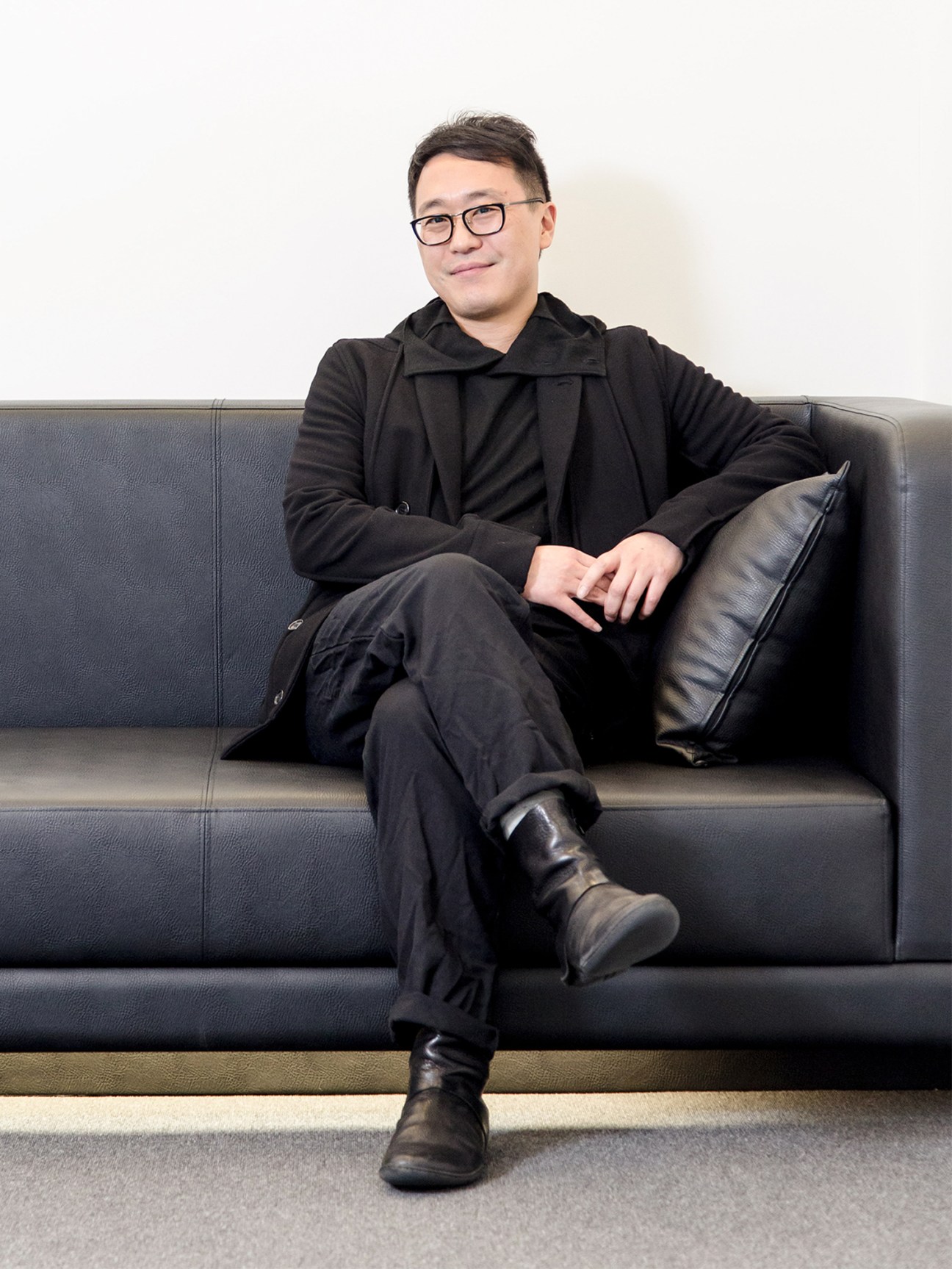 Doryun Chong, chief curator at M+ museum, sitting relaxed on a dark leather couch wearing glasses and black clothing