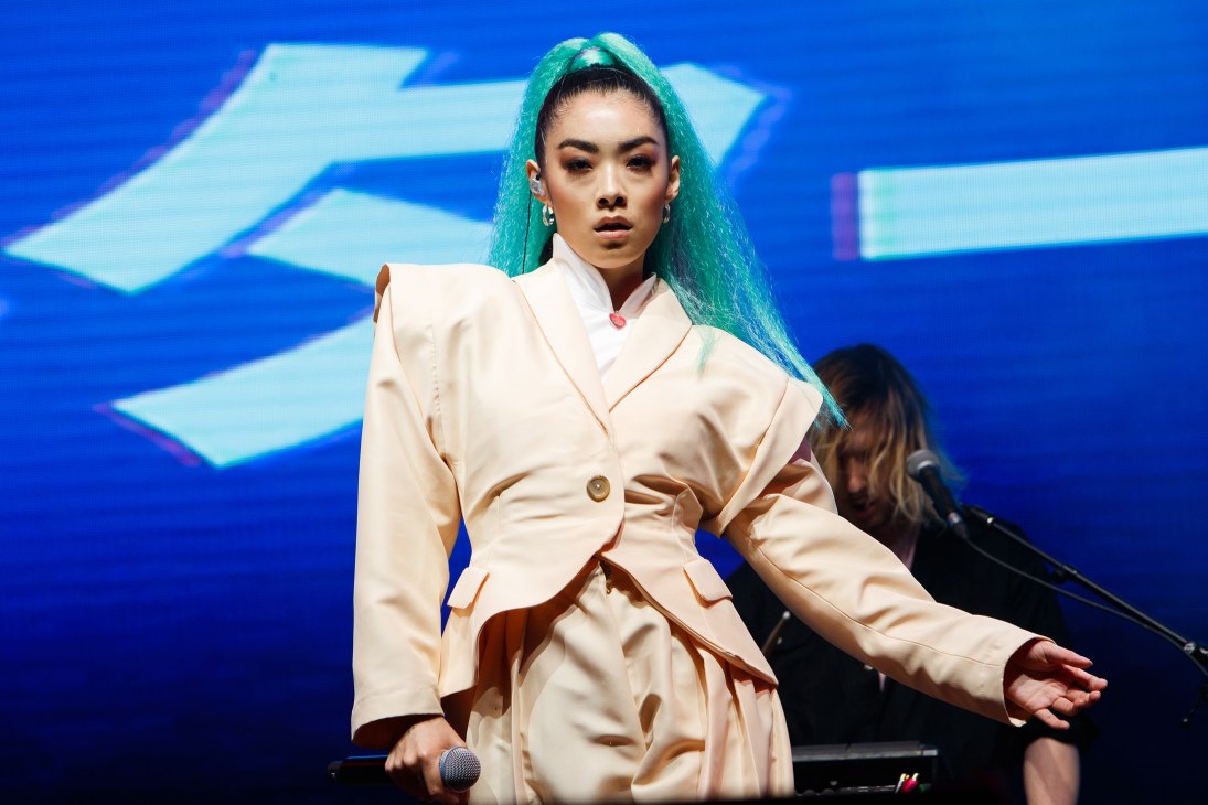 Rina Sawayama performing on stage in cream coat with turquoise hair against blue backdrop