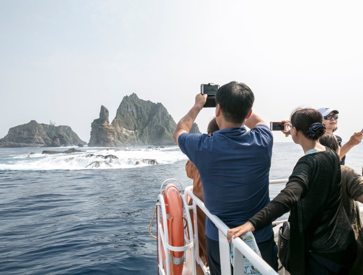 Tourists photograph dramatic rocky sea stacks from the deck of a boat on a sunny day.