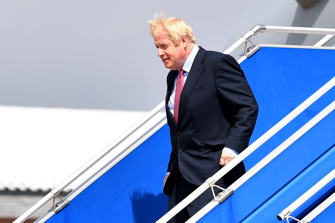 Boris Johnson walking down aircraft stairs wearing a dark suit and red tie