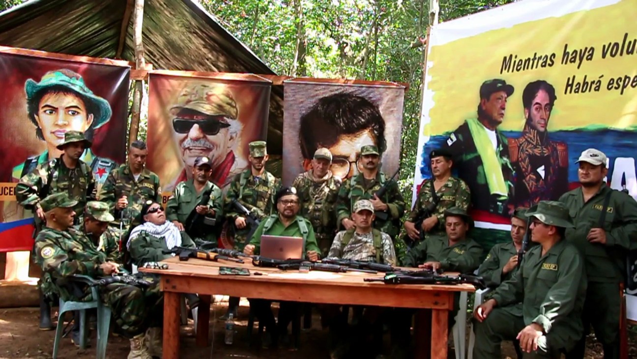 Armed men in military fatigues gathered around a table with portraits and banners in the background