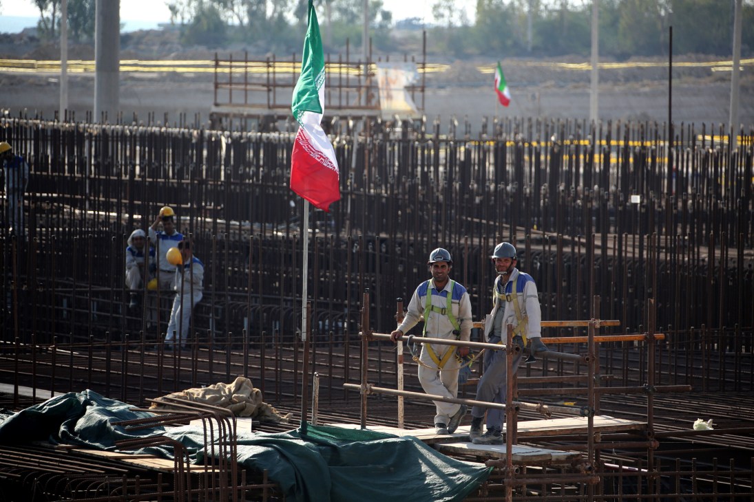 Construction workers at Bushehr nuclear power plant in Iran with Iranian flag flying over reinforced concrete structure.
