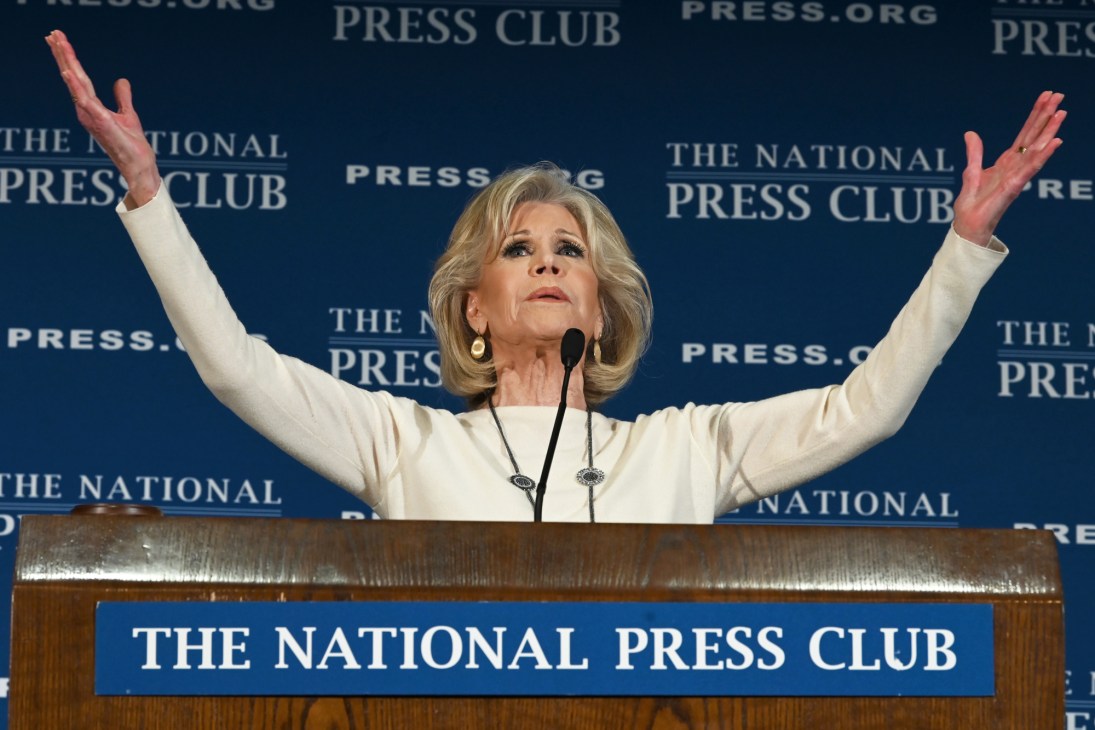 Woman in white blazer speaking at podium with arms raised at National Press Club event