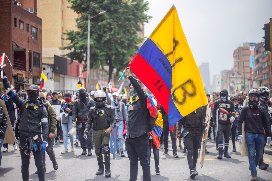 Protesters in dark clothing and face coverings march down a street carrying a large Colombian flag