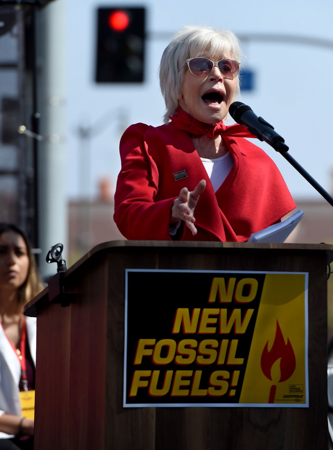 Jane Fonda speaking at outdoor rally wearing red jacket and sunglasses
