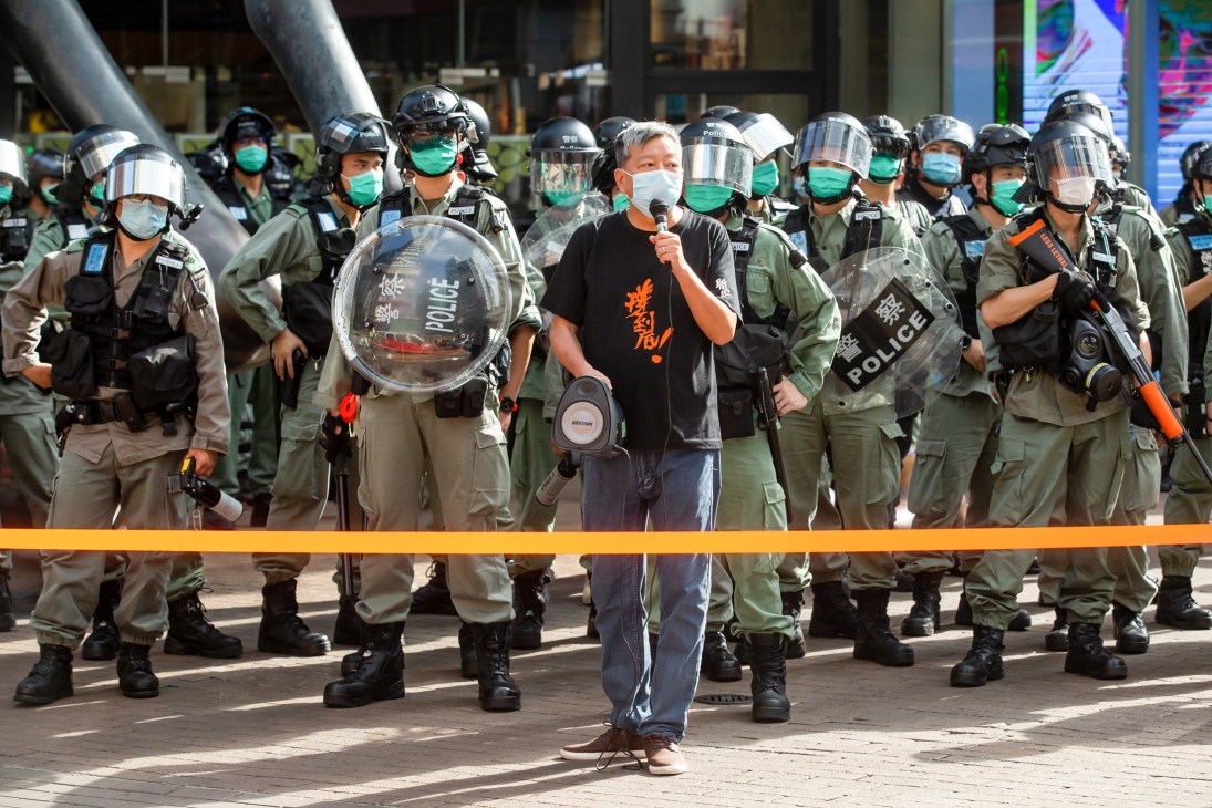 Riot police in protective gear and green uniforms stand behind a yellow barrier as a man in a black shirt holds a microphone during a Hong Kong protest.
