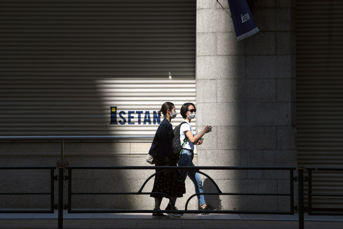 Two masked pedestrians walk past a closed storefront with shutters in Tokyo.