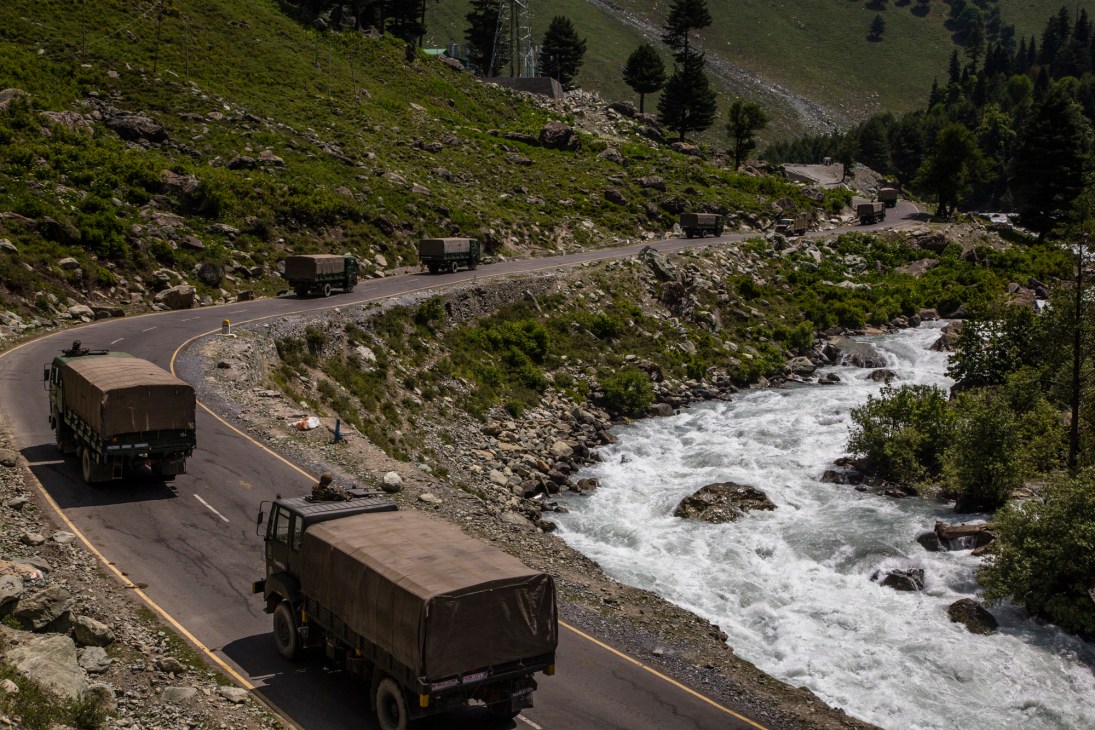 Military convoy of covered trucks traveling along a mountain road beside a rushing river