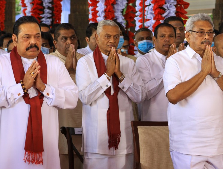 Three men in white clothing with red scarves performing prayer gesture with hands together