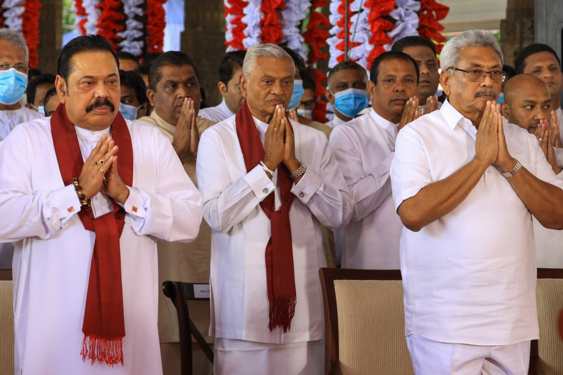 Three men in white clothing with red scarves performing prayer gesture with hands together