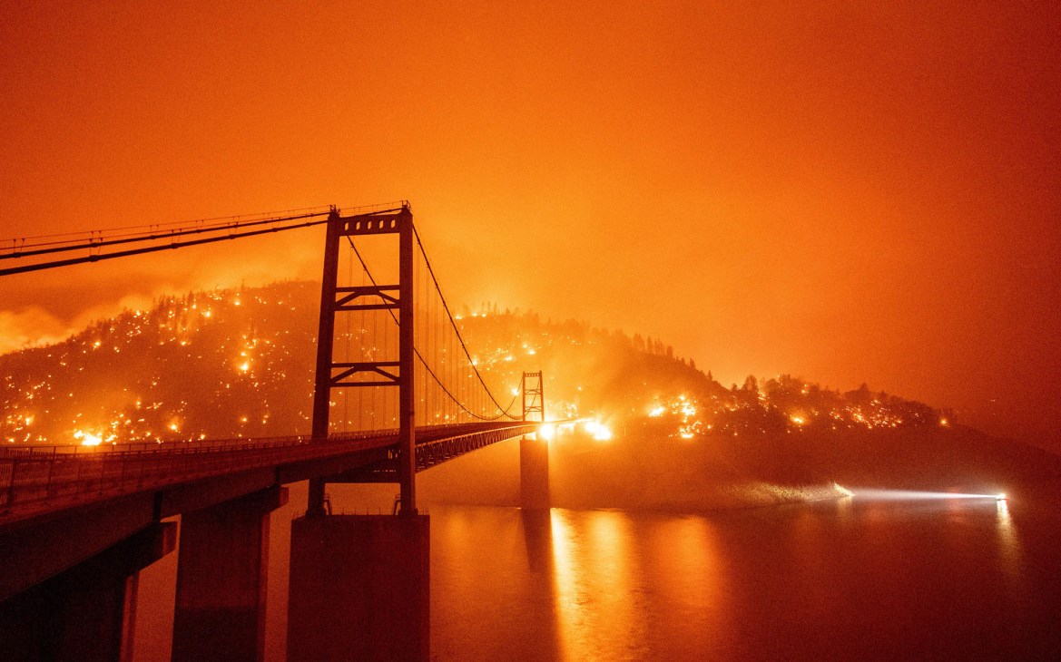 Bridge silhouetted against orange sky with wildfire flames visible on hills behind water