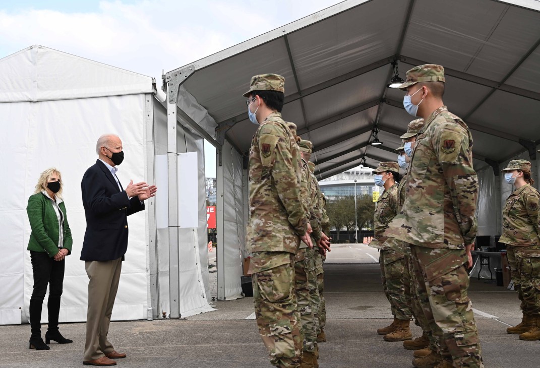 Biden and a woman in green speaking to military personnel in camouflage uniforms at a vaccination facility