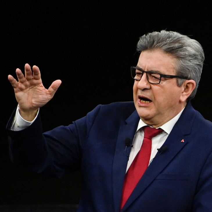 Man in navy suit and red tie gesturing with raised hand while speaking, wearing glasses against black background