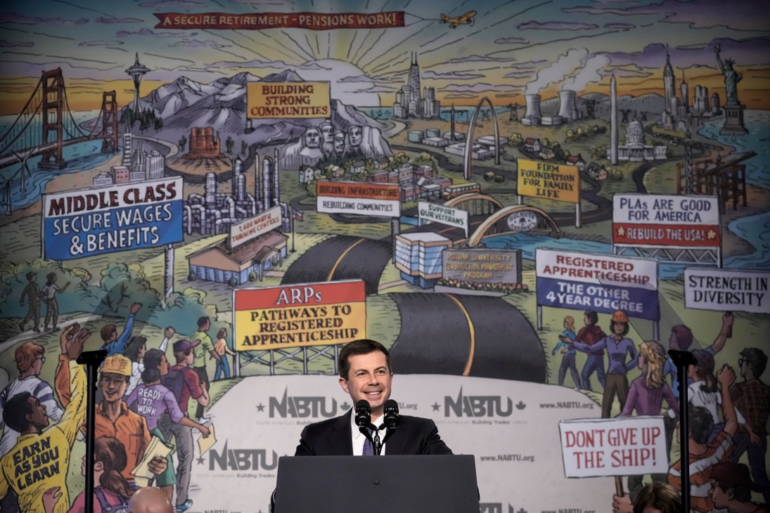 Transportation Secretary Pete Buttigieg speaking at podium with colorful mural backdrop featuring labor themes