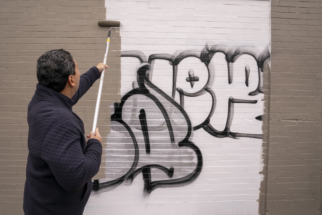 Person using paint roller to cover graffiti on brick wall with white paint