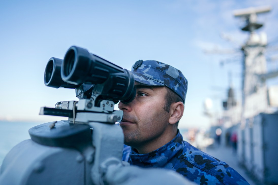 Naval officer using binoculars to observe from a ship with military vessel visible in background.