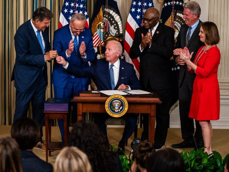 Joe Biden seated at presidential podium signing legislation while lawmakers applaud around him