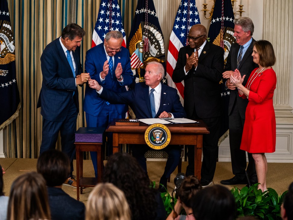 Joe Biden seated at presidential podium signing legislation while lawmakers applaud around him