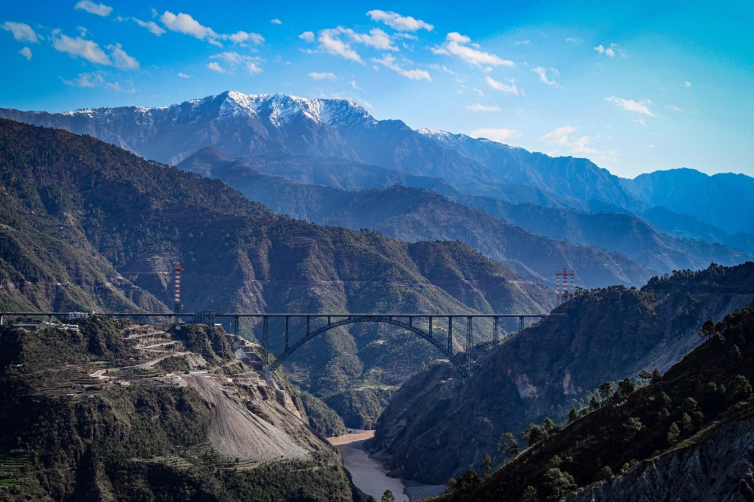 The Chenab Bridge under construction spanning a valley in Kashmir with snow-capped mountains in the background